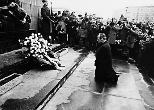 Willy Brandt kniet auf Stufen vor einem Blumenkranz. Im Hintergrund sind viele Zuschauer zu sehen
