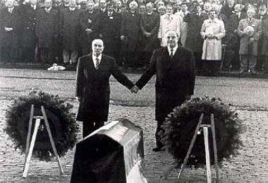 François Mitterrand und Helmut Kohl stehen vor zwei Blumenkränzen und halten die Hände. Im Hintergrund sieht man viele Zuschauer